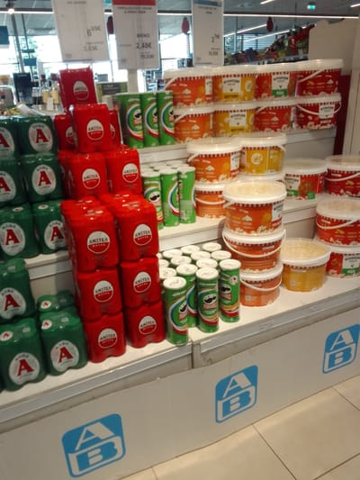 This media file captures a retail display of food and beverage products in a supermarket aisle. The shelves are stocked with a variety of items, including cans of what appear to be beverages with prominent "AMITEA" branding and green cans of Pringles chips. Alongside these are numerous tubs of popcorn in different flavors, such as "BUTTER," "PIZZA," and "BARBECUE," with "MYPOPCORN" branding.

Several price tags are visible, indicating prices in Euros. One tag shows "605€" with "3,03€" and "15,03€" below, suggesting a promotion or multi-buy offer. Another tag reads "379€" with "18,95€" beneath it. The background suggests an indoor retail environment with bright lighting and shelving units. The overall scene appears to be well-organized, typical of a grocery store display. The "A/B" logo, often associated with AB Vassilopoulos supermarkets in Greece, is visible on promotional cardboard barriers at the bottom of the image, confirming the location context. The image is a still shot with no discernable activity other than the static display of products.