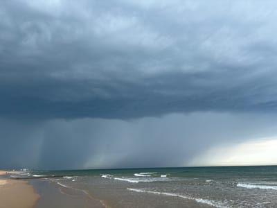The image depicts a dramatic coastal scene at Ostend, Belgium, under a turbulent sky. The foreground features a sandy beach meeting the gentle waves of the sea. In the distance, a breakwater runs parallel to the shore, and beyond that, several buildings and structures suggest a developed coastline. The sky is dominated by heavy, dark storm clouds, creating a somber and dramatic atmosphere. Visible rain streaks descend from the clouds in several areas, indicating an active weather event. The sea is a deep green, reflecting the overcast conditions, and the waves are moderate with whitecaps. A few faint figures can be seen walking on the beach to the left, appearing as small silhouettes against the vastness of the landscape. The lighting suggests it is daytime, likely late afternoon or early evening, given the intensity of the storm clouds and the muted light. The overall impression is one of impending or ongoing stormy weather at a coastal resort. There is no visible text in the image.