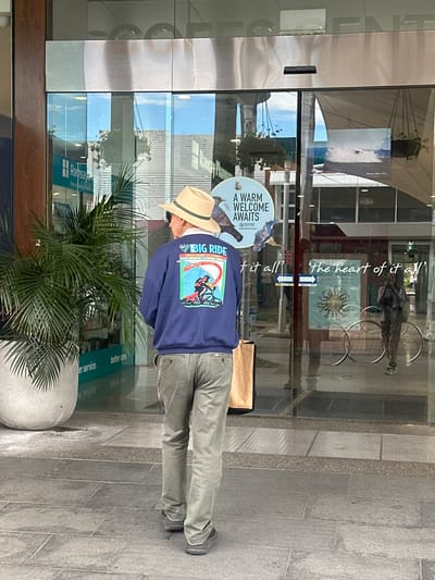 A man wearing a hat and a navy blue sweatshirt stands facing a set of glass doors. The sweatshirt has a graphic on the back that says "BIG RIDE" with text below it. The man is wearing khaki pants and dark shoes. He is standing on a tiled walkway. The glass doors reflect the surroundings, including plants, buildings, and people. A sign on the door reads "A WARM WELCOME AWAITS". Another sign on the door has text that says "'The heart of it all'". A woman is visible through the glass, taking a photo with a phone. The scene appears to be in a commercial area. The presence of "Ramsay Pharmacy" signage visible through the glass suggests the location is within the Coffs Harbour Health Campus in Coffs Harbour, Australia.