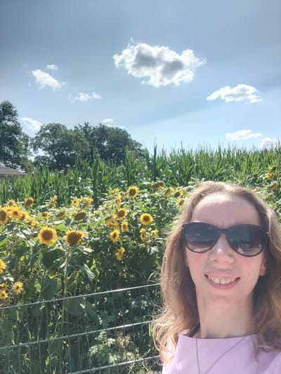 A woman wearing sunglasses and a pink shirt stands in front of a field of sunflowers and corn. The sky is blue with white clouds. The scene appears to be a rural area in Winterswijk Huppel, Netherlands, given the presence of agricultural crops and the natural landscape. The woman is looking at the camera and smiling.