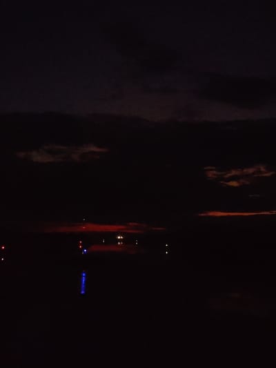 The image shows a night scene, likely in Bessonovo, Russia, given the prompt's information. The sky is dark with some cloud cover, possibly illuminated by a faint reddish glow on the horizon, suggesting either a sunset/sunrise or light pollution. In the lower portion of the image, there are several small points of light, some reflecting off a dark, still body of water. The lights appear to be various colors, including red, white, and blue, possibly indicating buildings or infrastructure along the shore. The overall scene is very dark, with low visibility.