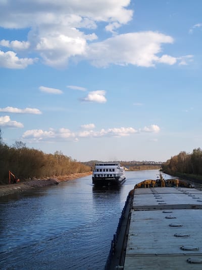 The image depicts a canal in Varvarino, Russia. In the foreground, the deck of a barge with multiple hatch covers is visible. A passenger ship is approaching in the canal. The banks of the canal are lined with trees in early spring foliage, and construction equipment is present on the left bank. A bridge and buildings can be seen in the distance. The sky is blue with scattered white clouds. The water in the canal is calm, with slight ripples visible.