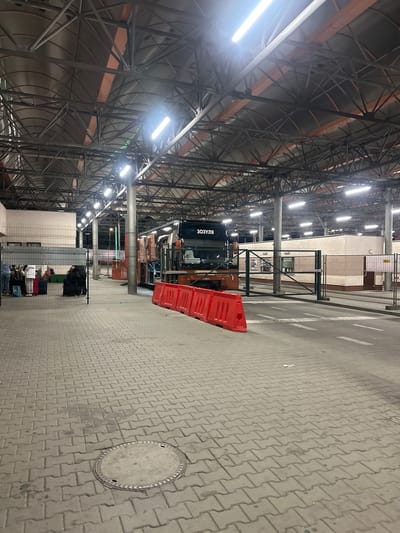 An outdoor nighttime scene depicts a bus station in Hrebenne, Poland, characterized by a covered waiting area with a metal truss roof illuminated by fluorescent lights. A large bus with the number "303" and text that reads "ЗОЗУЛЯ" is parked in the station. In the foreground, red plastic barriers line a road with white markings. The ground is paved with interlocking gray bricks, and a manhole cover is visible. On the left side of the image, behind a metal fence, several individuals with luggage are waiting. The overall environment appears functional and utilitarian, typical of a transportation hub.