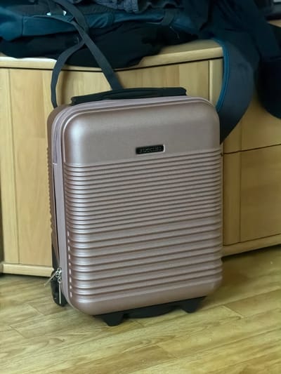 The image shows a rose gold carry-on suitcase with the "PUCCINI" logo on the front, resting on a wooden floor. The suitcase is positioned in front of a wooden cabinet. Above the suitcase, on top of the cabinet, are several dark-colored items of clothing, including what appears to be a backpack or duffel bag with straps. The scene is indoors, likely in a residential setting within Idar-Oberstein, Germany. There are no people visible in the image.