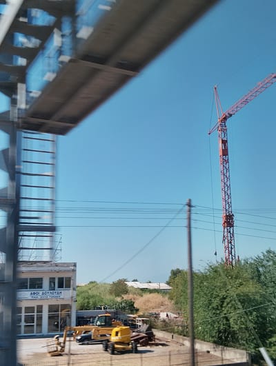 The image captures a construction site under a clear blue sky in the Municipal Unit of Echedoros, Greece. In the foreground, a blurred section of a modern building with glass windows is visible on the left. Further back, a prominent red tower crane stands tall against the sky. Various construction vehicles, including yellow excavators and a forklift, are parked on the site. In the background, a row of green trees lines the area, with some dry brush and distant buildings. The overall scene suggests ongoing construction or industrial activity.