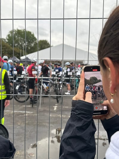 The image depicts an outdoor event scene in Weymouth, United Kingdom, observed through a metal barrier fence. In the foreground, the back of an individual's head, right ear, and right arm are visible. This person has dark hair, a small gold hoop earring with a white pearl drop, and is wearing a dark, textured jacket. They are holding a light-colored smartphone horizontally with both hands, actively recording a scene. The phone's screen displays a live video recording interface with a "00:00:03" timestamp, showing a cropped view of the event beyond the fence. The individual's fingernails are painted dark green.
Beyond the fence, a gathering of approximately 20-30 individuals, primarily adults, is visible. Many are equipped with cycling helmets and athletic wear, including colorful jerseys and at least one high-visibility yellow vest on the far left. Bicycles are arranged in rows, each with a visible number attached, suggesting a transition area for a triathlon or a cycling race. In the mid-ground, a large white marquee or tent stands prominently. Mature trees are visible in the background, beneath an overcast sky. The ground surfaces in the foreground and within the event area are damp, featuring several discernible puddles, indicating wet conditions.