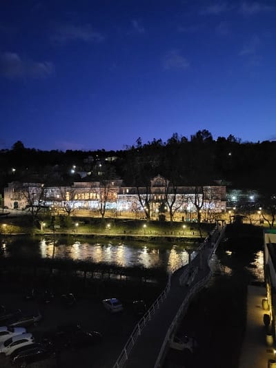 A tranquil night scene showcases a river reflecting soft lights from buildings lining its bank. Trees are silhouetted against the sky, while a bridge spans the river. Cars are faintly visible on the right side. The sky transitions from a deep blue above to darkness near the horizon. The scene evokes a peaceful, urban-meets-nature atmosphere.