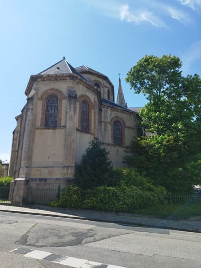 The image shows a view of a church from a street. The church building is made of light-colored stone and has a grey tiled roof. It features arched stained-glass windows and a tall spire topped with a cross. Lush green trees and bushes surround the church, with a black metal fence separating the property from the street. The street itself is paved and appears to be slightly sloped, with a manhole cover visible on the road. The sky is a clear, bright blue with a few wispy clouds, suggesting it is daytime on a pleasant day. The scene appears to be in a town or city, given the presence of the church and paved street, and the location context indicates Cherbourg-en-Cotentin, France. There are no people visible in the image. The overall impression is one of a serene, historic religious building set within a green, well-maintained environment.