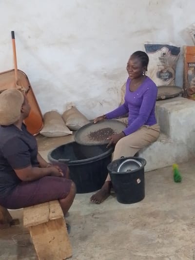 In Olomoda, Nigeria, an indoor scene depicts two women engaged in a processing activity. The woman on the right, wearing a purple long-sleeved top and light brown trousers, sits barefoot on a low concrete ledge. She holds a large, flat, circular metal sieve containing dark granular material over a black plastic tub on the floor. Her expression is a smile directed towards the woman on the left. The woman on the left, wearing a dark short-sleeved top and a dark maroon skirt, is seated on a wooden bench, facing the first woman. Her head is covered with a light brown wrap. Between them, on the floor, is the black plastic tub, and a smaller black bucket containing a metal bowl is near the first woman's feet. The background consists of a plain, light-colored wall. Against the wall, a wheelbarrow is visible on the left, several burlap sacks are on the floor, and two large white sacks labeled "DAGOTE BREAD FLOUR" and another with "BOM" are stacked on the right. A green plastic bottle lies on the floor to the far right. The concrete floor shows minor scuffs and marks.