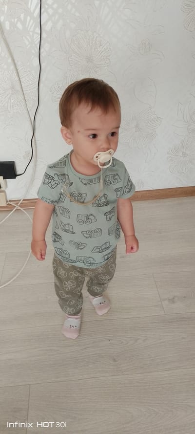 A full-body, eye-level shot shows a young Caucasian child, appearing to be a toddler, standing on a light-colored wooden floor. The child has light brown hair and is wearing a light blue t-shirt with a repeating pattern of various construction vehicles, and khaki-colored pants with a leaf pattern. The child also has on light pink socks with a white animal face design on the front. A white pacifier is in the child's mouth, attached by a clear and beige string to the t-shirt. The child is looking off to the right of the frame. Behind the child is a white wall with a textured floral pattern. A black electrical cord hangs vertically on the left side of the wall, connected to a black electrical adapter. The environment suggests an indoor setting, possibly a home. There is no indication of the child's specific location within the city of Kedrovy, Russia.