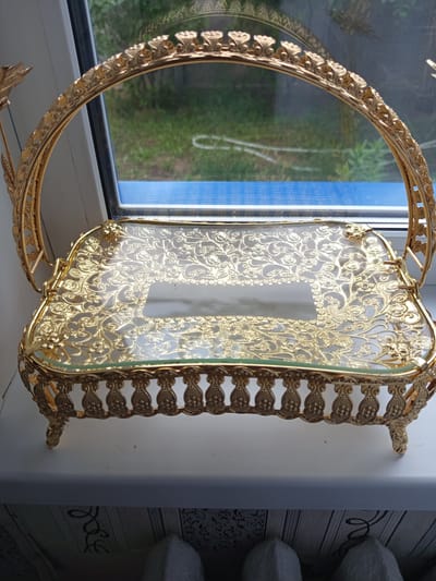 This media file shows a golden ornate serving tray with a glass surface. The tray has intricate metalwork featuring floral and vine motifs, and it rests on four decorative legs. A curved, arched handle made of the same golden metal is attached to the sides of the tray. The tray is positioned on a white windowsill, with a blue band of light visible below it, suggesting it is placed in front of a window. Outside the window, green foliage and grass can be seen, indicating an outdoor setting with natural light. The overall impression is of an elegant decorative item, possibly used for serving food or as a centerpiece. There is no visible text in the image.