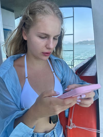 In Nam Ninh Hoa, Vietnam, a young woman with blonde hair is on a boat, looking down at her pink smartphone. She is wearing a white bikini top and a light blue unbuttoned shirt, with denim shorts. The background shows the ocean and distant islands. A life preserver is visible on the boat.