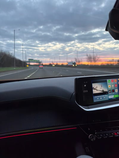 A front passenger perspective view inside a vehicle traveling on a multi-lane highway near Garden City, United Kingdom, at dusk or dawn. The sky overhead is dominated by dense, textured grey and white clouds, transitioning to a prominent band of orange and red along the horizon, indicative of sunset or sunrise.
The vehicle's interior features a dark dashboard with a textured carbon fiber pattern on the lower section and a red ambient light strip running horizontally across it. A modern infotainment screen is prominently displayed on the central-right side, showing a digital clock indicating "23:5," an ambient temperature of "7°C," and a media interface. The media displayed is a video featuring a person on a surfboard or wakeboard over water, with accompanying text "J Balvin, Ryan Castro, DJ Snake - Taki" visible below. Application icons for phone, music, maps, messages, settings, YouTube, Netflix, TikTok Live, and Auto/CarPlay are also visible on the screen. Below the infotainment screen, physical controls and buttons are partially visible. A glimpse of a leg in grey trousers is seen at the very bottom right, indicating an occupant.
Outside the vehicle, the highway stretches into the distance with multiple lanes marked by white lines. Tall grey streetlights line the left side of the road. A blurred green road sign is visible in the distance. Ahead, a white passenger car with illuminated red taillights travels in a central lane. Further down the highway on the right, a large white truck with bright headlights or running lights is in motion. Sparse, dark trees and bushes line the road verges and the distant horizon. The road surface appears damp in places.
