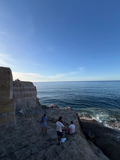 The image depicts a sunlit coastal scene in Sperone, Italy, showcasing a rugged rock formation bordering the sea under a clear blue sky with minimal high-altitude cloud cover.
The sky, occupying the upper portion of the frame, transitions from a lighter blue near the horizon on the left to a deeper azure towards the top right. The sun, positioned low on the left, casts a warm glow, creating some backlighting effects.
The Mediterranean Sea dominates the middle and right sections of the image, appearing as a deep blue body of water with discernible ripples and white-capped waves crashing against the rocky coastline in the lower right.
A large, multi-layered cliff, composed of light brown and gray sedimentary rock, rises on the left side of the image. Its sheer face and horizontal strata are clearly visible. Several individuals are situated on the flat top of this cliff, mostly silhouetted, appearing to be observing the view or resting.
Below the main cliff and extending towards the water, a series of darker, irregular rock formations and platforms create a natural terraced landscape. On one of these lower platforms, three individuals are present:
1. A person in a blue and white striped t-shirt, light blue shorts, and flip-flops, holding a red and white striped towel, stands facing away from the cliff and towards the sea.
2. To their right, a person in a white t-shirt and dark shorts is seen from the back, holding a white garment.
3. Next to them, a third person in a white t-shirt and pink or red shorts is seated on the rock, also viewed from the back.
Near these individuals, a white plastic cup and a white plastic bag or piece of litter are visible on the rocks.
Further down and to the right, a person with a backpack is seen navigating or climbing the steep, dark rocks near the water's edge. At the lowest accessible rocky platform, directly adjacent to the waves, two individuals are lying or sitting on a blue towel, appearing to be sunbathing or relaxing. The overall scene suggests a leisurely gathering in a natural coastal environment.