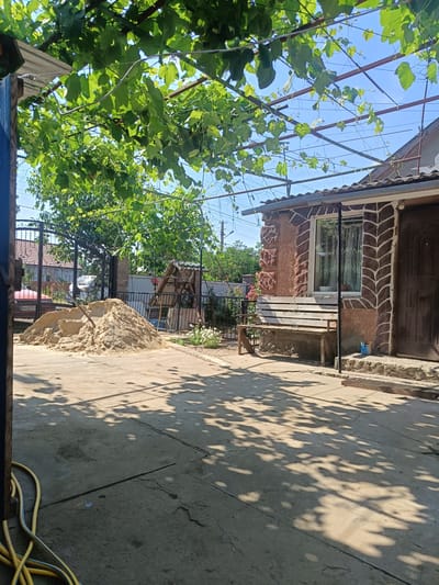 This media file depicts a sunny outdoor scene, likely in a rural or suburban backyard. The foreground is dominated by a concrete patio with a yellow hose coiled on the left side, partially in shadow. A large pile of light-colored sand sits in the middle ground, with a shovel stuck in it, suggesting ongoing construction or landscaping work.

Behind the sand pile, a black metal gate leads to a yard with a red car visible beyond it. To the right of the sand pile is a rustic wooden bench situated in front of a building. The building has a stone and stucco facade with a decorative pattern around the window and door. A dark wooden door is on the right side of the facade. The window has white curtains. A small blue can, possibly paint, sits on the step in front of the door.

Overhead, a dense canopy of green grape leaves, supported by a metal frame, casts dappled shadows on the patio, indicating a warm, sunny day. The sky is clear and blue. In the background, trees and other structures can be seen, hinting at a residential area. The overall impression is one of a domestic setting, perhaps undergoing some renovation or preparation for an outdoor project. The bright sunlight and shadows suggest the time of day is likely midday or early afternoon. No people are visible in the scene. No visible text is present. The location is specified as Peremozhne, Ukraine.