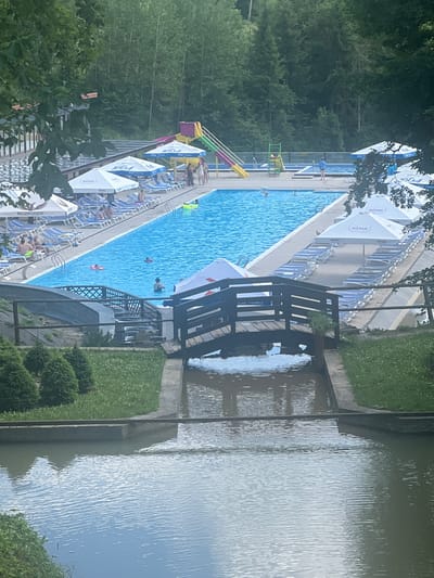 The image depicts a sunny day at a swimming pool complex, likely located in Khashchovania, Ukraine. The primary feature is a large, bright blue swimming pool filled with people. Several individuals are visible in the water, some swimming and others relaxing. In the foreground, a small wooden bridge spans a narrow waterway. Surrounding the pool, rows of empty lounge chairs are set up under large white umbrellas, suggesting a well-maintained recreational area. In the background, a vibrant waterslide complex with yellow and green slides adds to the playful atmosphere. Lush green trees and foliage frame the scene, indicating an outdoor setting. The overall impression is one of leisure and summer enjoyment.