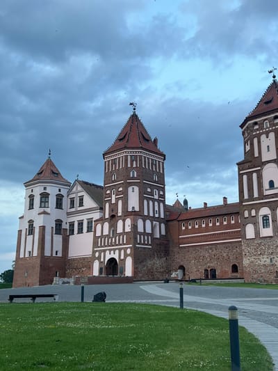 The image displays the Mir Castle Complex, located in Мірскі сельскі Савет, Belarus, under an overcast sky. The dominant structure is a multi-turreted castle constructed of red brick and varying stonework. Two prominent octagonal towers with pointed, red-tiled roofs, each topped with a dark weather vane, are visible. The central tower features white-plastered panels contrasting with its red brick facade, including arched recesses and a visible entrance archway at ground level. To the left, a smaller, lighter-colored tower with a conical red roof is connected to the main structure. Connecting wall sections, constructed from a mix of stone and brick, exhibit rectangular window openings and various architectural details.
In the foreground, a segment of green grass with sparse white flora occupies the lower-left. Adjacent to it, a light-colored, patterned paved courtyard or plaza extends towards the castle, featuring an curved edge. A dark-colored, slatted park bench is situated on the grass. Several dark, cylindrical bollards or pathway lights are evenly spaced along the border between the grass and the paved area. Two dark, round, cannon-like objects are present on the paved surface near the base of the castle's central tower. The sky is filled with layered grey and blue clouds. Numerous small, dark birds are observable in various positions, both in flight across the sky and perched on the castle's roofs and architectural elements. No human subjects are present within the scene.
