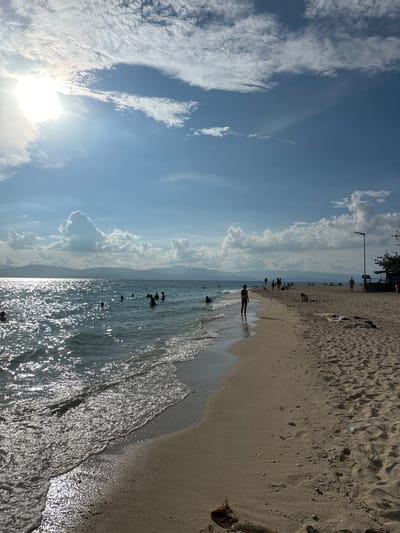 The image depicts a sunny day at a beach in Saavedra, Philippines, with the sun high in the sky and bright sunlight reflecting off the water. The sea is calm with small waves lapping at the sandy shore. Several people are visible in the water, swimming or wading, and on the beach, some are walking, sitting, or lying down. In the background, mountains form a distant silhouette against the horizon. The sky is partly cloudy with white cumulus clouds. A few structures, including a lamppost and what appears to be a small building or kiosk, are present on the right side of the beach. Debris, such as a piece of driftwood, is scattered on the sand.