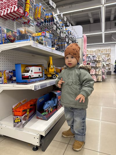 The image shows a young child standing in front of a shelf in a store, examining toys. The child is wearing a brown patterned hat, a green jacket with a fur-trimmed hood, blue jeans, and brown shoes. The shelf is stocked with various toys, including Hot Wheels cars and trucks, a toy ambulance, and a shark-themed toy. The location is likely a retail establishment in Korobovo, Russia, based on the presence of items and labels written in languages commonly found in that region. The child is looking at the camera with a slightly inquisitive expression. The environment appears to be a well-lit store aisle.