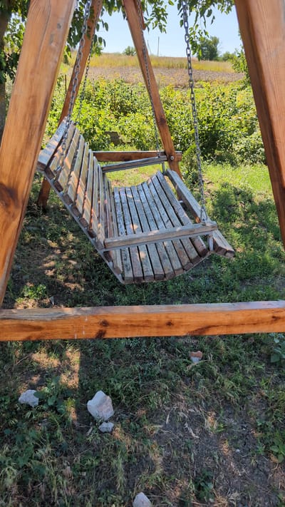 A wooden swing set with a slatted wooden seat is positioned in a grassy area. The swing seat is suspended by metal chains from the wooden A-frame structure. In the background, there is a field with green vegetation and some distant trees under a clear sky. The ground around the swing set is covered in grass and small stones. The image appears to be taken outdoors during daylight.