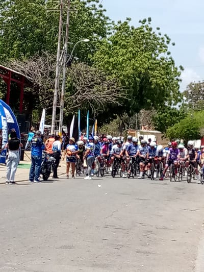 The image shows a group of cyclists lined up on a paved road, seemingly at the start of a race or event. They are wearing cycling gear, including helmets, and are mounted on their bicycles. Several people stand on the left side of the road, possibly organizers or spectators. There are banners and flags visible near the start line. A motorcycle is parked near the people on the left. Behind the group of cyclists are trees and buildings in the background. Based on information not in the image, the location is the city of Guatacaral, Venezuela.