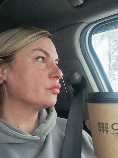 A woman in a gray hooded sweatshirt, wearing a seatbelt, is holding a brown paper coffee cup in a car. The cup is partially visible, displaying a wheat stalk logo. The woman is looking up with a slight smile, her blonde hair partially visible. A car window and interior elements form the background.