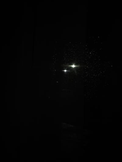 This image depicts a nighttime scene in the city of Griboyedov, Armenia, characterized by extreme darkness. Two prominent, horizontally aligned bright white light sources are visible in the upper right portion of the frame, likely originating from streetlights or distant vehicle headlights. The right side of the image, particularly surrounding these lights, is covered with numerous small, reflective points, consistent with water droplets on a transparent surface such as a window or windshield, suggesting rain or condensation. The remainder of the frame is obscured by profound darkness, rendering any specific environmental features, structures, people, or objects indistinguishable. No actions or interactions are discernible.