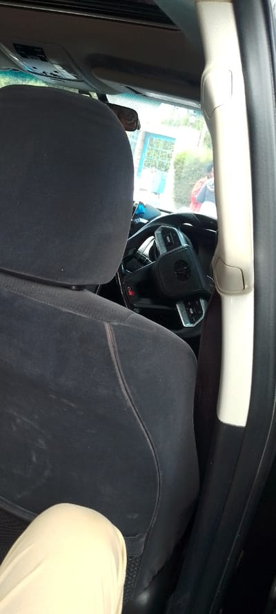 Inside a car, a black cloth headrest dominates the left view. A white interior car pillar stands tall on the right. A steering wheel with a prominent center emblem is visible through the gap. Outside, figures are seen near a blue-framed window. A tan pant leg appears in the lower left corner, adding a layer to the composition.