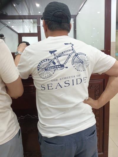 An individual wearing a white t-shirt and a black baseball cap stands with their back to the camera. The t-shirt features a graphic of a bicycle with words arranged in a circular pattern around it, and the words "THE STREETS OF SEASIDE" printed below. The individual is standing in what appears to be an indoor setting, possibly a shop or market, with wooden paneling visible in the background. There is another person partially visible to the left of the frame, also wearing a white shirt.