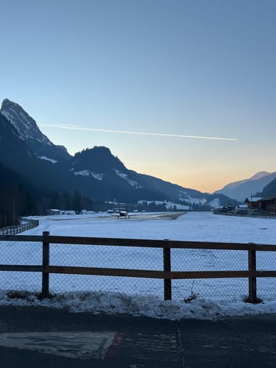 This image depicts a winter scene at Saanen Airport (LSGK) in Saanen, Switzerland, likely during sunrise or sunset.
In the foreground, a dark asphalt surface is partially covered by a thin layer of snow and slush. A faded white and red ground marking, possibly an airfield symbol, is visible on the asphalt. Immediately behind this, a wooden-post and chain-link fence, partially covered in snow, spans the width of the frame. A snowdrift is present behind the fence.
The midground features a snow-covered airfield. A cleared asphalt runway or taxiway extends from the left-center towards the distant right. A dark-colored, single-engine propeller aircraft, possibly a turboprop, is positioned on this paved surface, facing generally away from the viewer. To the left, a curved wooden fence runs along the edge of the snow-covered field. Several small, utilitarian buildings, consistent with airport infrastructure, are visible on both sides of the airfield, particularly to the right.
The background is dominated by snow-capped mountains and coniferous forests. A prominent, jagged peak is visible on the far left. The sky transitions from clear blue overhead to a pale orange-yellow gradient near the horizon, indicative of a low sun angle. Two distinct white contrails are present in the upper sky, one longer and more prominent than the other. No people are visible in the scene.