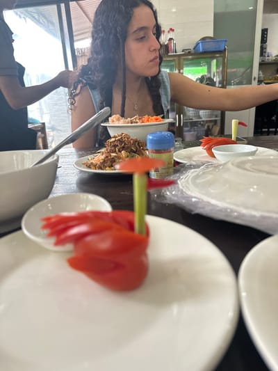 A woman with dark, curly hair sits at a table set with plates. Some plates are stacked with plastic between them. A plate holds food resembling a salad. Other plates are adorned with tomato slices. A metal serving spoon rests on the table. A display case with shelves is visible behind the woman, and natural light enters from a window to the side.