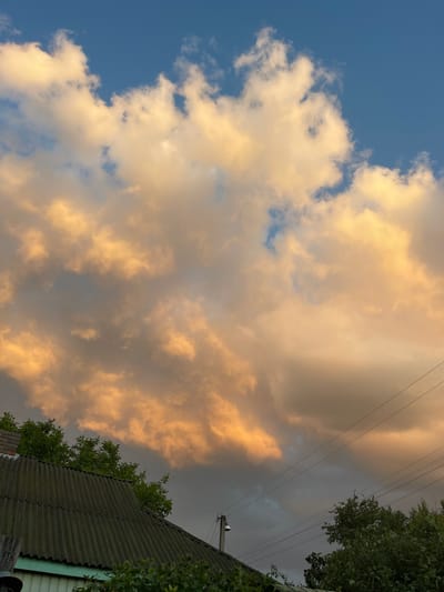 The image depicts a sky filled with dramatic, golden-hued clouds, likely illuminated by the setting sun. In the foreground, the corrugated roof of a building is visible, with green foliage and trees surrounding it. Telephone wires traverse the lower portion of the sky. The overall scene suggests an outdoor environment, possibly a rural or semi-urban area, with a focus on the atmospheric conditions and the interplay of light and shadow on the landscape. The location is specified as Krasnokutsk, Ukraine.