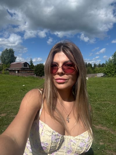 A young woman with blonde hair and pink-tinted sunglasses takes a selfie in a grassy field. She is wearing a yellow, floral-patterned top and a necklace with a cross pendant. Behind her, a wooden fence encloses a field with a rustic wooden house and trees. The sky is blue with scattered white clouds. The location is identified as Pastukhovo, Russia. A white animal, possibly a dog, is visible in the distance.