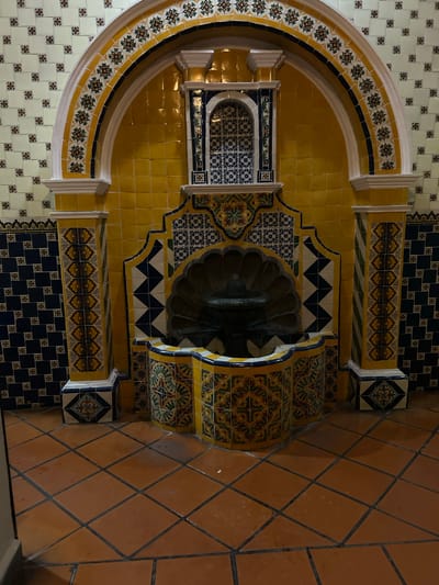The image depicts a tiled water fountain in Santa Ana Yancuitlalpan (Chilhuacán), Mexico. The fountain is set against a wall covered in white and floral patterned tiles. A large, arched structure surrounds the fountain, decorated with yellow, white, and floral patterned tiles. The fountain itself is constructed with yellow and blue tiles, featuring a scalloped basin. The floor is covered with brown, diamond-patterned tiles.
