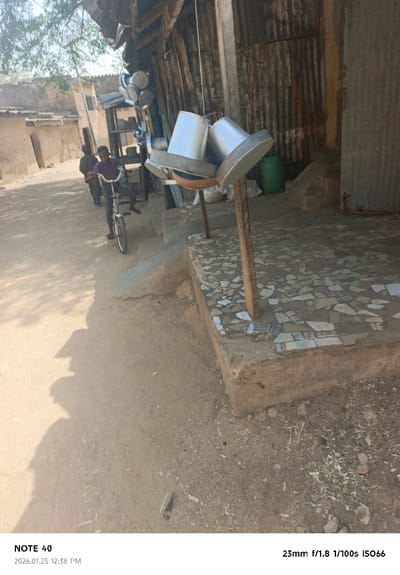 This image depicts a sunlit outdoor scene in Tudun Wazurchi, Nigeria, captured on 2026.01.25 at 12:38 PM.

The foreground consists of a dirt ground path, extending from the lower left to the upper center. On the right, a raised concrete platform, approximately 0.5 meters high, is visible. This platform is surfaced with irregularly shaped fragments of light-colored ceramic tiles. Two inverted, silver-colored metal pots with flanged edges are displayed on a rustic wooden stand positioned on the tiled platform. Behind this, a corrugated metal building wall, exhibiting rust and weathered appearance, is visible. Adjacent to the wall, a green plastic barrel and a white sack are present on the ground. Further back on the right, a multi-tiered wooden or metal shelf holds several smaller silver-colored pots or bowls.

On the dirt path, two individuals are present. In the mid-ground, a young male, appearing to be a child or adolescent, wears a purple long-sleeved shirt and dark trousers. He is astride a dark-framed bicycle, with his feet not touching the pedals. He faces generally towards the viewer's left. Behind him, another individual, dressed in dark clothing, walks or stands further down the path, partially obscured by the first person.

The left side of the frame features sunlit, rudimentary buildings constructed from earth-toned materials, likely mud or unpainted plaster, with flat roofs. Sparse tree foliage is visible in the upper left corner. Strong shadows, including an elongated shadow consistent with a standing human figure (likely the photographer) and other objects, are cast prominently on the dirt path in the left foreground, indicating bright overhead sunlight. The overall environment suggests a rural or semi-urban street setting.