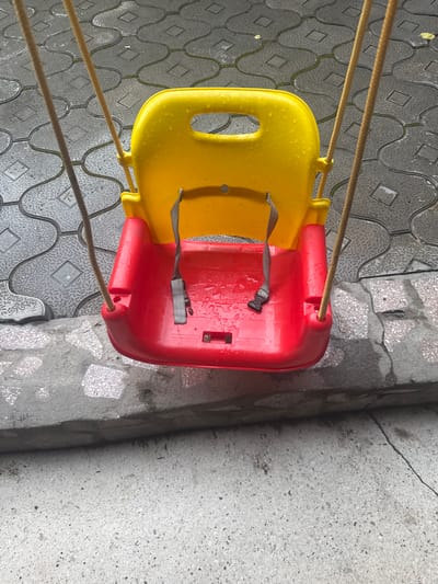 The image shows a yellow and red plastic baby swing suspended by brown ropes. The swing is wet, suggesting it has been raining. It is positioned over a textured grey pavement, possibly outdoors. To the right of the pavement is a rougher, grey surface that appears to be concrete. The swing has two grey safety straps with black buckles. The scene is likely set in a residential area given the materials of the ground and the presence of a swing. The lighting suggests it is daytime, and the wetness on the swing and pavement points to recent rain. There is no text visible in the image.