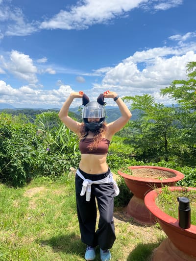 A woman with curly brown hair stands in front of a lush, green hillside with a blue sky and scattered clouds above. She is wearing a brown tube top, black pants tied with a white shirt, and light blue sneakers. A black motorcycle helmet with brown fuzzy ear attachments is on her head. She is holding her hands up, forming heart shapes above the helmet. Two large, terracotta planters are visible in the foreground, one with dry soil and the other with sparse green growth. A black water bottle rests on the edge of the planter with greenery. The woman is facing forward and smiling, with mountains and trees visible in the background. The scene appears to be a scenic overlook in Sơn Điền, Vietnam.