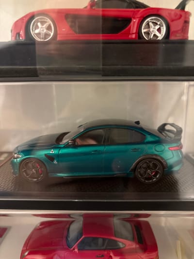 The image displays three vertically stacked, clear display cases, each containing a die-cast model car. The specific location within Kajang Municipal Council, Malaysia, cannot be determined from the visual information provided.

The uppermost case, partially visible, houses a red model sports car, identified as a Mazda RX-7 (FD3S) variant, positioned facing right. It features chrome five-spoke wheels.

The central case prominently displays a dark metallic teal/green model of an Alfa Romeo Giulia Quadrifoglio sedan, viewed from its left side and oriented towards the right. This model possesses a black roof, a black rear spoiler, and black multi-spoke wheels with visible red brake calipers. It rests on a textured black base inside the transparent enclosure.

The lowermost case, also partially visible, contains a red model sports car, identifiable as a Porsche 911 (993 generation) variant, also viewed from its left side. A prominent red rear wing is visible.

No individuals are present in the scene. The ambient lighting produces reflections on the clear surfaces of the display cases.
