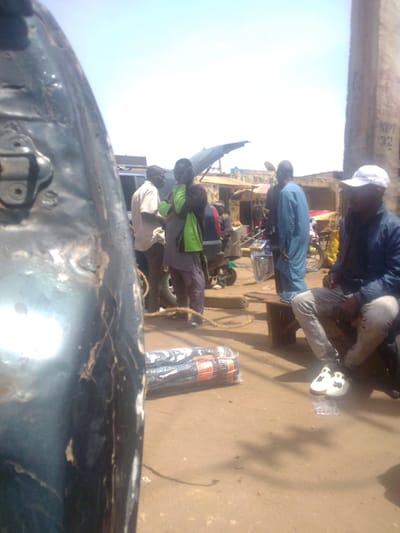 The scene depicts an outdoor urban environment, identified as Mista Ali, Nigeria, under bright daylight. The foreground on the left is dominated by a dark, metallic surface, likely part of a vehicle, exhibiting wear and a visible latch mechanism. The ground is unpaved, consisting of light brown earth with visible shadows.
In the mid-ground, a group of approximately eight adult males are present. On the left, three men stand: one in a light shirt and dark trousers, facing right; another in a dark long-sleeved garment with a bright green vest/scarf, facing left; and a third partially obscured. Near them, a length of light brown rope is coiled on the ground. Two rolls of dark material, wrapped in clear plastic and secured with orange/red tape, rest on the ground in the central foreground.
Further back, a dark SUV-type vehicle with its rear hatch open is parked. To the right of the standing group, a man in a light blue traditional garment stands, facing left. Behind him, a motorbike is parked. On the far right, a man in a dark blue jacket, light jeans, and a white baseball cap is seated on a low wooden bench, looking right. He wears white sneakers with black accents and appears to be holding a dark object in his lap. Another motorbike is partially visible in the background on the right.
The background features several single-story structures, consistent with market stalls or small shops, some with corrugated metal roofs and visible signage. A tall, light-colored vertical structure with "NIPP 32" visible text stands on the far right. The sky is clear and bright blue.