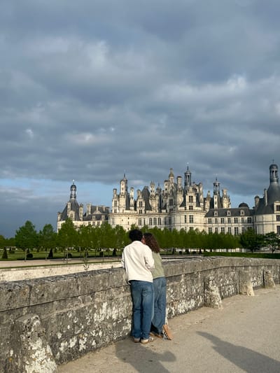 The image depicts a couple standing on a stone bridge in front of the Château de Chambord, located in Chambord, France, at coordinates 47.616 latitude, 1.514 longitude. The couple is facing the castle, with the man on the left, wearing a white jacket and blue jeans, and the woman on the right, wearing a brown shirt and blue jeans. The castle is a large, white structure with many towers and spires. The sky is cloudy with a slight grey tinge. The ground around the bridge is covered in gravel. A low stone wall lines the bridge. The bridge is covered in moss and is surrounded by trees. The setting appears to be a sunny day with a slight breeze. The scene suggests a romantic and picturesque location.