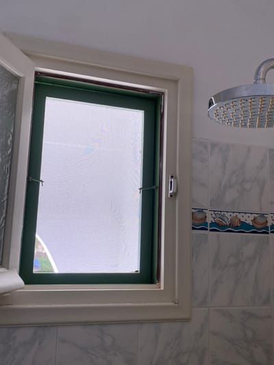This image depicts a bathroom interior in Πλατύς Γιαλός, Greece, during daytime. A cream-colored window frame is centrally positioned, containing a closed dark green-framed insect screen secured by two small, metallic pivot latches. The screen diffuses the external light, rendering the outside view indistinct but bright. A partial view of a textured glass pane is visible to the immediate left of the main window frame. To the right of the window, the wall is tiled with light grey, marble-patterned ceramic tiles. A horizontal decorative border featuring blue waves and stylized seashells (conch, scallop, clam) is visible above these tiles. A circular, chrome-plated shower head with numerous spray nozzles is mounted on the wall in the upper right quadrant of the image. No individuals or actions are present.