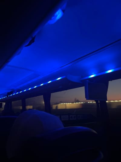 The image shows the interior of a bus, lit by blue LED lights running along the top of the aisle. The bus appears to be moving, as the scene outside the windows blurs. The image is taken from the perspective of a passenger seated in the bus, looking up at the blue lights and toward the front of the bus. The passenger is wearing a white cap. The location of the image is Goyencourt, France, based on the coordinates provided. The scene is a dark, narrow space, illuminated only by the blue lights and the outside world seen through the windows.