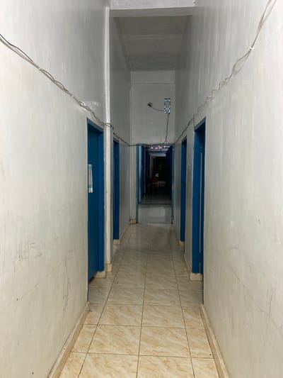 This image depicts a narrow, well-lit hallway within a building in Tanjung Gading, Indonesia. The hallway is lined with several identical blue doors on either side, suggesting a multi-room dwelling or establishment. The walls are painted a light, off-white color and show signs of age and minor scuff marks. Electrical wires are visible running along the top of the walls, secured by white clips. The floor is tiled with light-colored, marble-patterned tiles. At the far end of the hallway, a slightly darker area is visible, with what appear to be red and white triangular flags hanging. The scene is devoid of any people or overt activity, presenting a static, observational view of the interior space. The overall impression is one of a functional, perhaps modest, corridor within a residential or commercial building in the specified Indonesian location.