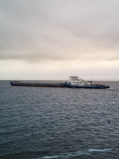 The image shows a tugboat pushing a barge on a body of water. The sky is overcast. The tugboat is painted with a blue and white color scheme. The barge is long and low to the water. The location is Prislon', Russia. There are no people visible in the image.