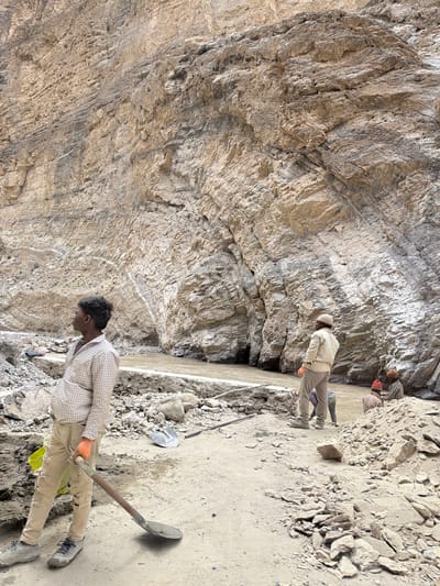 In Stongde, India, a group of men are working in a rocky, mountainous area beside a muddy river. The terrain is rough, with loose stones and debris scattered everywhere. A large, imposing cliff face dominates the background, its striated layers of rock suggesting geological history. In the foreground, a young man wearing a plaid shirt, ripped trousers, and orange gloves stands with a shovel, looking off to the left. Further back, another man in a light-colored jacket and cap stands near the riverbank, facing away from the camera. Two other individuals, one with a red hat, are seated or crouched by the river. The overall impression is one of manual labor in a rugged natural environment.