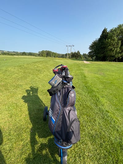 The image shows a golf bag and cart on a golf course at The Pocklington Golf Club, located in Pocklington, United Kingdom, at latitude 53.938 and longitude -0.757. The golf bag is black with blue accents, and it has a number of clubs inside. The cart is on the fairway, facing towards a grove of trees in the distance. The ground is covered in green grass. The sky is blue and clear, with a few wispy clouds. The sun is shining brightly. There are no people visible in the image. The course is in good condition and there are power lines in the background.