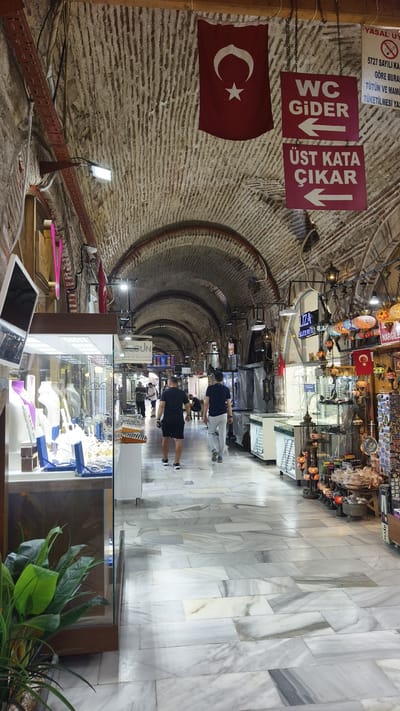 The image depicts the interior of a covered bazaar, likely a market in İzmir, Turkey, given the Turkish flag and signage. The architecture features a long, arched passageway with a stone ceiling, illuminated by artificial lights and hanging lamps. The floor is covered in large, grey and white marble tiles.

On the left side, a shop displays jewelry and watches in glass cases, with a potted plant in the foreground. Further down the passageway, other shops are visible, some with merchandise displayed outside. In the center of the frame, two men are walking away from the camera, one in a black t-shirt and shorts, the other in a dark blue t-shirt and light-colored pants. Several other people are seen in the distance, walking or browsing in the shops.

Hanging from the ceiling are several signs. A Turkish flag is prominently displayed near the top. Two rectangular signs, one red with white lettering, point to the left. The top sign reads "WC GIDER" (WC goes), and the bottom sign reads "ÜST KATA ÇIKAR" (Go to the upper floor). A larger, white sign with red lettering is also visible on the right, containing Turkish text and a prohibition symbol. The shops on the right appear to sell decorative items, including colorful mosaic lamps. The overall impression is a bustling marketplace with a traditional architectural style.