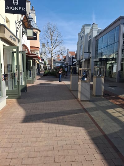 The image displays a pedestrian shopping street within Ingolstadt Village, Ingolstadt, Germany. The scene is illuminated by direct sunlight under a clear blue sky, casting distinct shadows across the light brown rectangular paver walkway.
On the left, a multi-story building prominently features a black "AIGNER" sign. Multiple storefronts on the ground floor exhibit open, light green-framed glass doors and window displays. Adjacent to this, a building with a conical turret and a "nite" sign is visible.
On the right side of the walkway, three freestanding outdoor display cases are arranged in a line. Each unit consists of a transparent glass top section showcasing merchandise, including handbags and shoes, atop a light grey rectangular pedestal. "BOSS" branding is visible within at least two of these cases. Beyond these displays, a modern building with a large glass facade reflects the surroundings, and further down the street, storefronts for "GUCCI" and "WOOLRICH" are visible.
In the mid-ground, a single male individual stands to the right of the center. He has short, dark hair and is dressed in a dark t-shirt and blue jeans. He is observed holding a smartphone with both hands and appears to be actively interacting with it, looking down at the screen. Behind him, a leafless deciduous tree stands centrally, indicating a non-summer season. The overall environment is an outdoor commercial retail area.