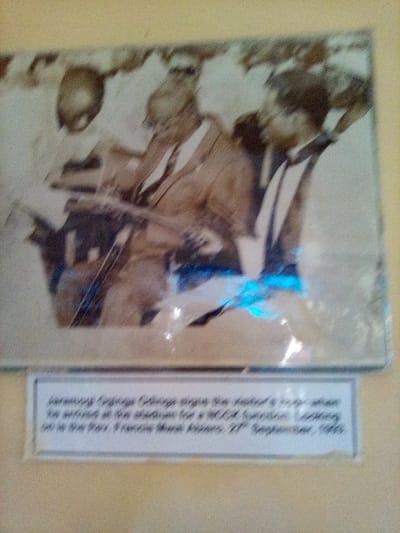 A framed, sepia-toned or monochrome photograph is displayed on a light yellow wall in South Sakwa ward, Kenya. The image within the frame is partially obscured by blur and reflections, notably a blue glare in the lower central and right portions. The photograph depicts three male figures prominently. In the center, an older man identified as Jaramogi Oginga Odinga, with a beard and wearing a light-colored suit jacket, white shirt, and dark tie, is actively signing a document or logbook with a pen. His left hand appears to steady the document, while his right hand holds the writing instrument. To his right, observing the action, stands a man identified as Rev. Francis Mwao Adwera, wearing a white shirt and a dark tie. A third individual, wearing a light shirt, is partially visible to Odinga's left. The scene appears to be an outdoor or semi-outdoor public gathering, possibly at a stadium, with indistinct figures in the background. A white label placed below the photograph provides the context: "Jaramogi Oginga Odinga signs the visitor's log when he arrived at the stadium for a RCCK function. Looking on is the Rev. Francis Mwao Adwera, 27th September, 1993."