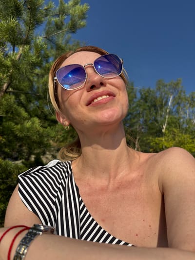 A woman is looking up at the sky, wearing sunglasses and a striped top. She is outdoors, with trees and a blue sky in the background. The image appears to be taken in Shuiskaya Chupa, Russia.